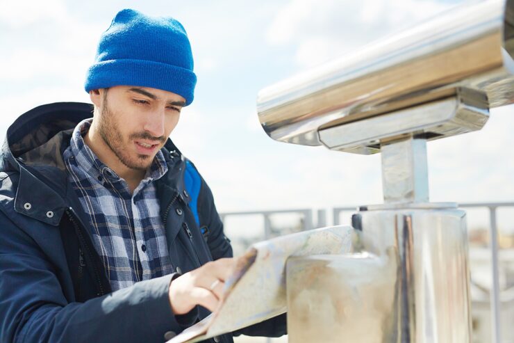 A man wearing a blue toque and a plaid shirt underneath a dark jacket, intently looking at and holding a folded map or checklist next to a metal sightseeing viewer, possibly representing a tourist or homeowner reviewing maintenance plans.