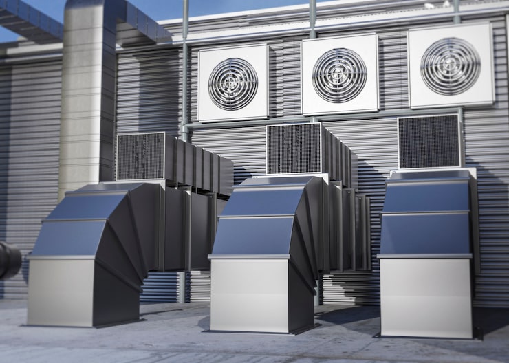 A large, industrial exterior view of multiple commercial HVAC units on a rooftop, including condenser fans and prominent, angular sheet metal ductwork.