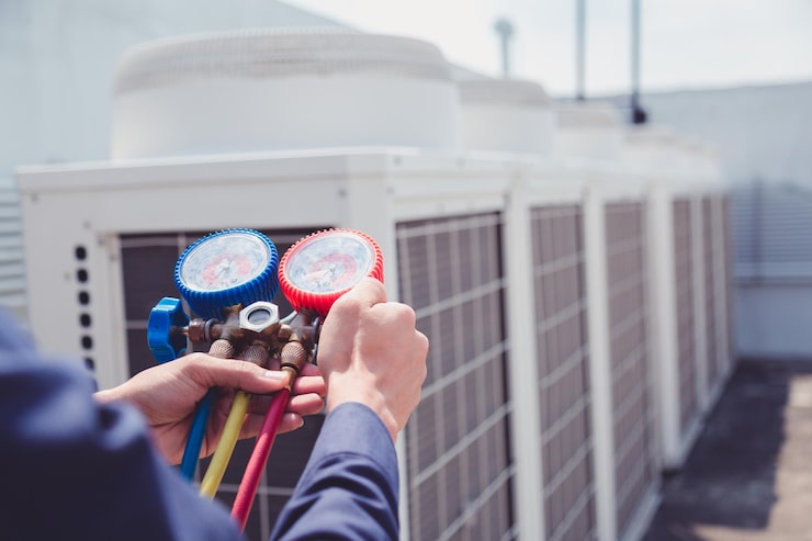 A close-up view of a technician's hands holding a manifold gauge set with red, blue, and yellow hoses, used for checking refrigerant pressure on outdoor commercial HVAC units blurred in the background.