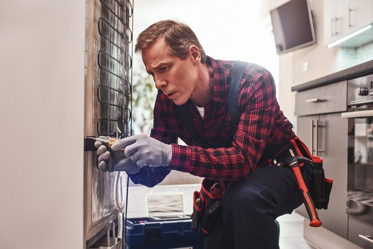 A repair technician in a red plaid shirt and work gloves, focused on fixing the coils on the back of a large household appliance, possibly a refrigerator or freezer, which serves as a general illustration of an appliance repair worker.