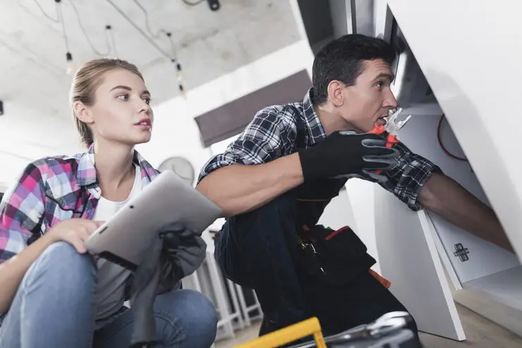 A woman holding a tablet observes a male HVAC technician as he inspects an indoor component.