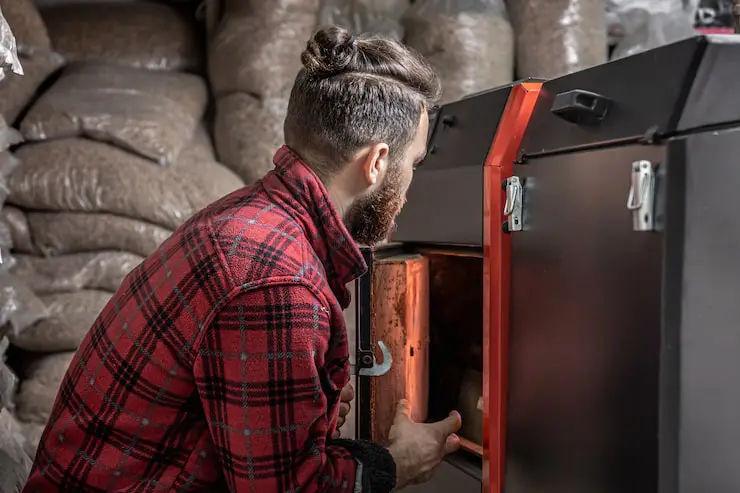 Man in plaid shirt checking the combustion chamber of a furnace, surrounded by wood pellets.