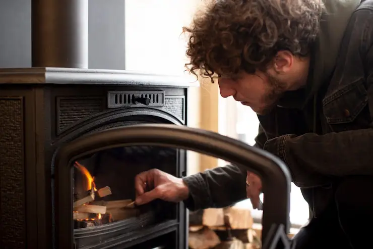 Man with curly hair crouches to add small pieces of wood to a fire in a black wood stove.
