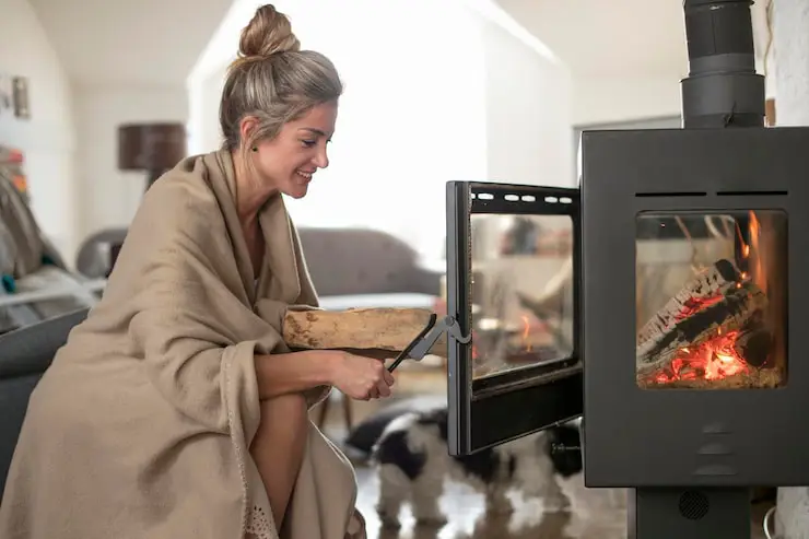 Smiling woman wrapped in a blanket adds wood to a modern, glass-front wood-burning stove.